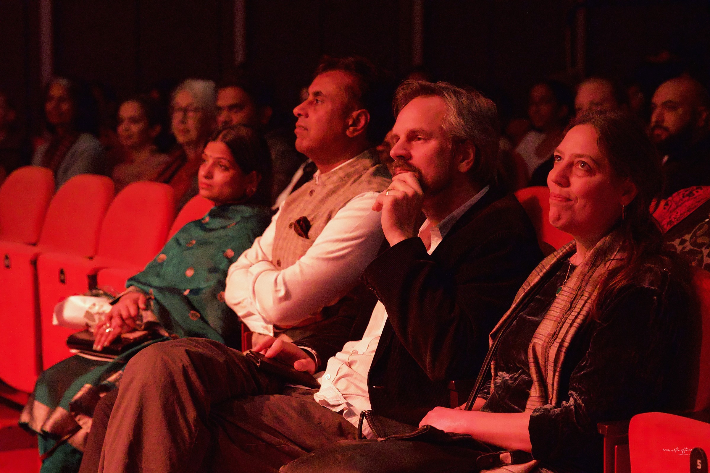 Four audience members seated together, watching a performance, lit in red stage light