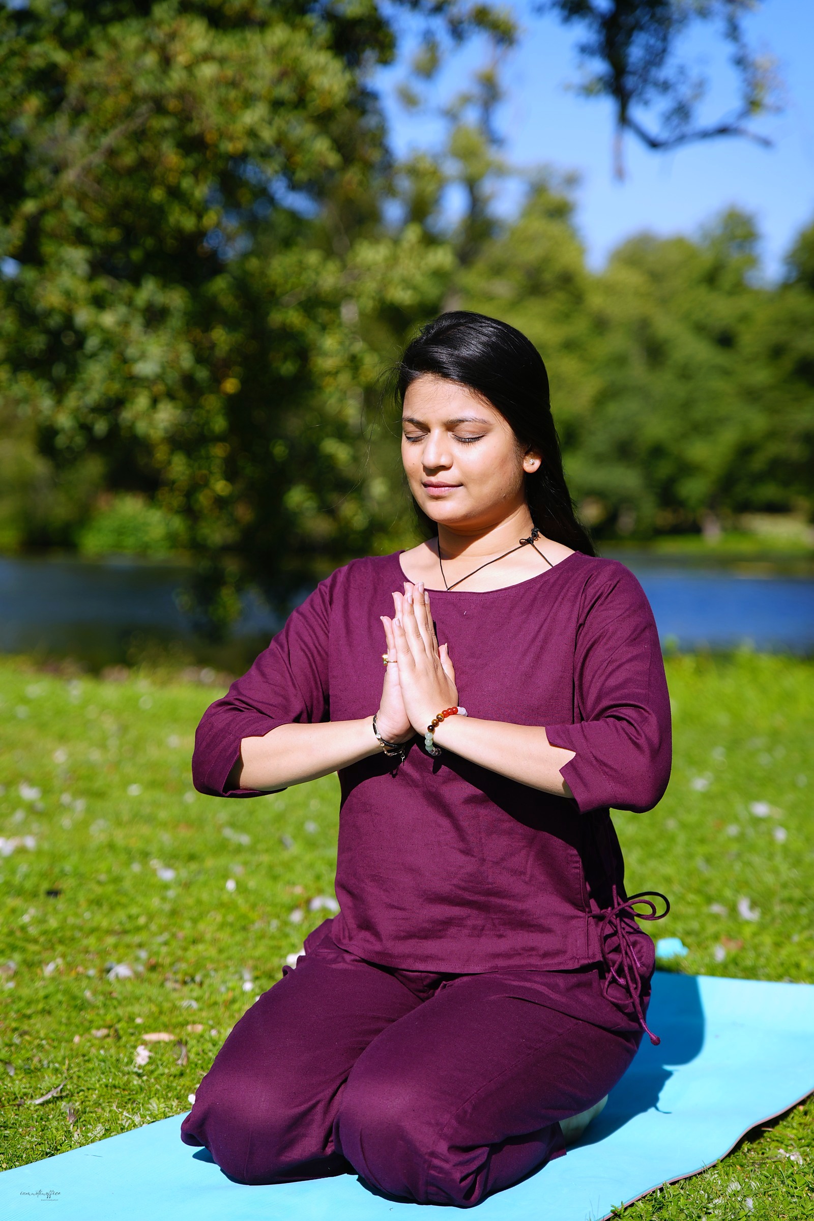 Woman seated in prayer hands pose with eyes closed, sunlit park background